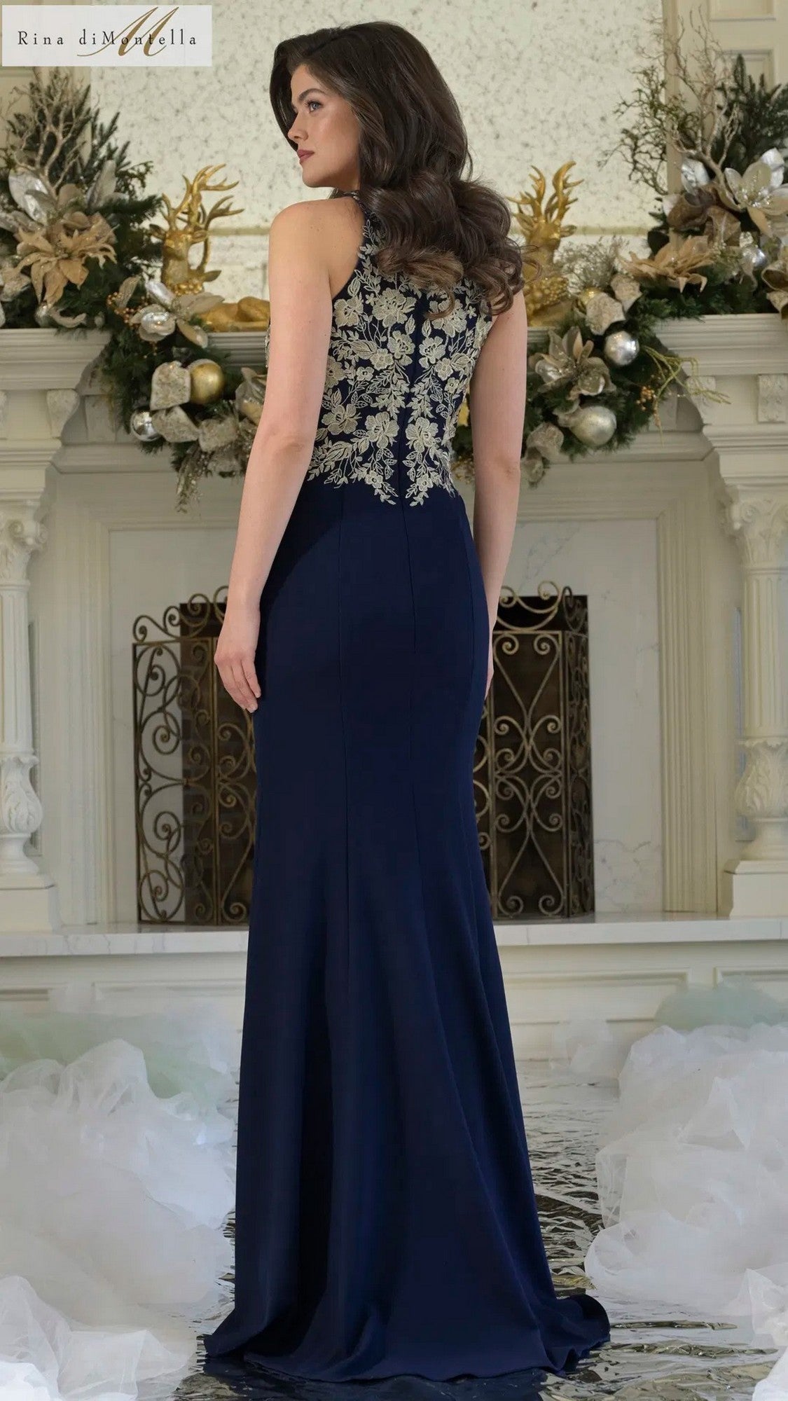 A woman wearing the Rina Di Montella RD3345 long formal dress in navy stretch crepe with gold floral embroidery on the back stands before a decorated white fireplace.